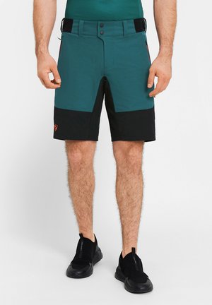 Mann trägt farblich abgesetzte kurze Hosen in Türkis und Schwarz mit schwarzen Slip-on-Sportschuhen, steht vor einem schlichten weißen Hintergrund.