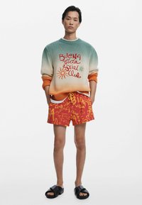 Sweter z dzianiny w gradientowych odcieniach zieleni do pomarańczu, z grafikami. Kontrastowe szorty bawełniane w kolorze czerwonym z żółtym napisem. Czarne klapki.