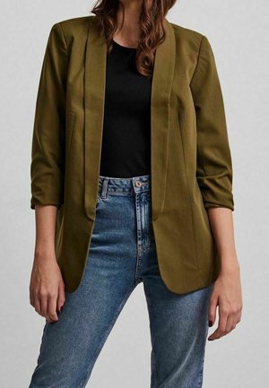 Woman wearing an olive green blazer with 3/4 sleeves over a black top and high-waisted blue jeans, standing against a neutral background.