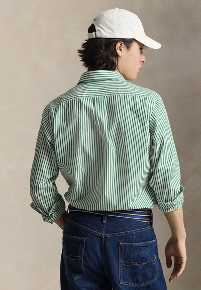 Homme vu de dos portant une chemise rayée verte et blanche avec les manches retroussées, un jean bleu, une ceinture rayée et une casquette de baseball blanche.