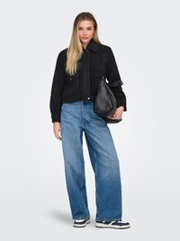 Jeune femme aux longs cheveux blonds, portant une veste noire, un jean large bleu, des baskets et tenant un grand sac à main noir.