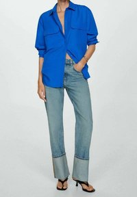Camicia blu oversize con bottoni e due tasche sul petto, abbinata a jeans chiari a zampa d'elefante con dettagli sui lati e sandali neri con tacco.
