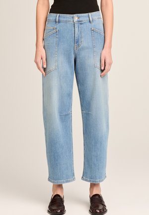 Hellblaue Jeans mit geradem Bein und großen vorderen aufgesetzten Taschen, kombiniert mit einem schwarzen Oberteil und dunklen Slip-on-Loafers.