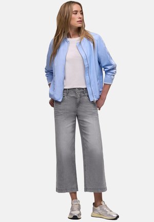 Femme debout, les mains dans les poches, portant une veste bomber bleu clair, un T-shirt blanc, un jean gris à jambes larges et des baskets blanches.