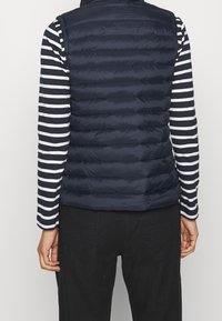 Tmavě modrá proš ívaná vesta se zakládáním do vodorovných pruhů, nošená přes černo-bílé pruhované tričko s dlouhým rukávem. Má vysoký límec a zavírání na zip.