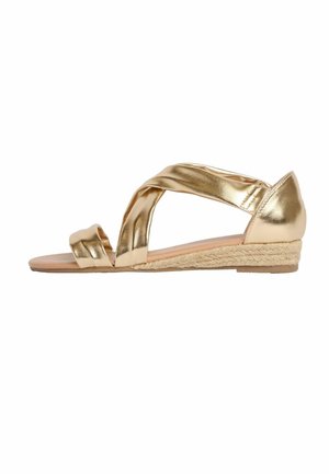 Gouden metallic lage sleehak sandaal met gekruiste bandjes en open teen op een gevlochten jute zool, gezien van opzij tegen een witte achtergrond.