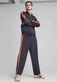 Fato de treino azul-marinho com padrão e detalhes laranja, composto por um casaco com fecho éclair e punhos elásticos, combinado com ténis de cor clara.