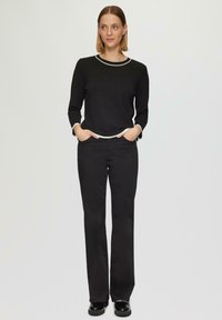 Zwarte long sleeve top met witte rand aan de halslijn en manchetten, gecombineerd met zwarte rechte broek. Schoenen zijn zwart met een glanzende afwerking.