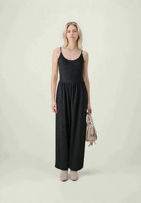 Black sleeveless jumpsuit with a fitted bodice and wide leg design, made of textured fabric. Paired with a light-colored handbag and shoes.