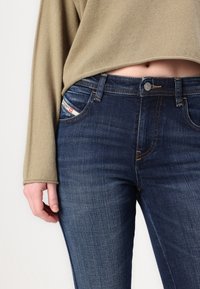 Vaqueros de mezclilla en azul oscuro con sutiles desgastes y costuras naranjas. Un bolsillo frontal presenta una etiqueta de la marca; combinado con una blusa de punto beige.