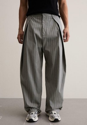 Čovjek nosi sive široke hlače na pruge s naborima, uparene sa sivim i bijelim tenisicama, stoji na jednobojnoj pozadini.