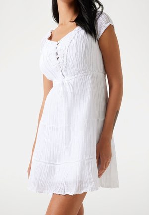 Frau trägt ein weißes, knielanges Kleid mit kurzen Ärmeln, strukturiertem Stoff, Spitzenbesatz am Ausschnitt und einem Taillengürtel.