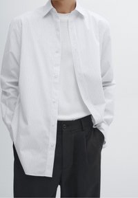 Chemise blanche à rayures avec col, portée sur un t-shirt blanc uni, associée à un pantalon noir ajusté.