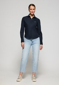 Camisa de botones azul marino con un pequeño logo, combinada con jeans rectos azul claro y zapatillas beige. Tejido suave, cuello clásico.