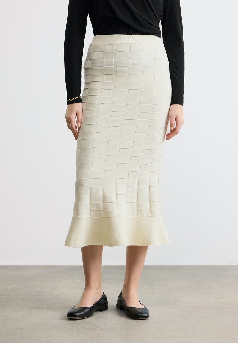 Femme portant une jupe midi texturée crème avec un ourlet évasé, un haut à manches longues noir, des ballerines noires et un bracelet en or au poignet gauche.