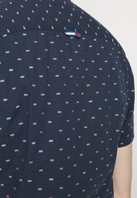 Tmavě modrá košile s krátkým rukávem a vzorovanou texturou, zdobená malými růžovými, bílými a modrými puntíky. Na zadní straně je malý trojbarevný proužek.