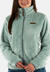 Chaqueta de forro polar de color azul claro con un patrón texturizado, cuello alto, cremallera completa en el frente y un pequeño parche de logo marrón en el pecho.