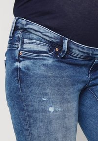Vaqueros de denim en azul medio con un lavado deslavado, cinco bolsillos, remaches de cobre y un ligero desgaste en la zona del muslo.
