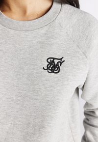 Grijze trui met een geribde ronde hals en een zwart geborduurd logo op de linkerborst. De stof heeft een zachte textuur en een casual pasvorm.