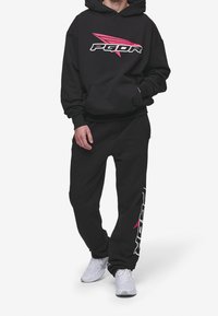 Černá sada mikiny s kapucí a tepláků z měkké látky. Mikina má velké bílo-růžové logo PGDR; na kalhotách je detail loga po straně.