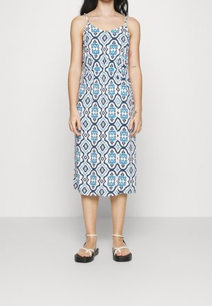 Kobieta w sukience bez rękawów, sięgającej kolan, z niebiesko-białym geometrycznym wzorem oraz białych sandałach na paski, stojąca na tle jednolitego tła.