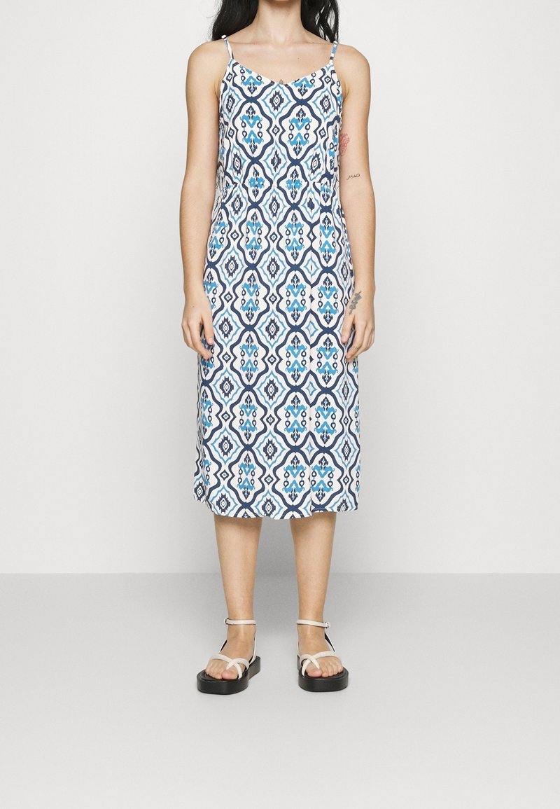 Femme portant une robe sans manches, longueur genou, avec des motifs géométriques bleus et blancs, et des sandales blanches à lanières, debout devant un fond uni.