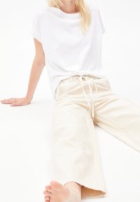 Femme assise par terre portant un t-shirt blanc et un pantalon large crème avec une taille à cordon, pieds nus sur fond blanc.