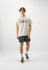 T-shirt alb Nike cu text "Bulls" și mâneci crem, asortat cu pantaloni scurți albaștri cu un model și adidași negri. Șosete albe vizibile.