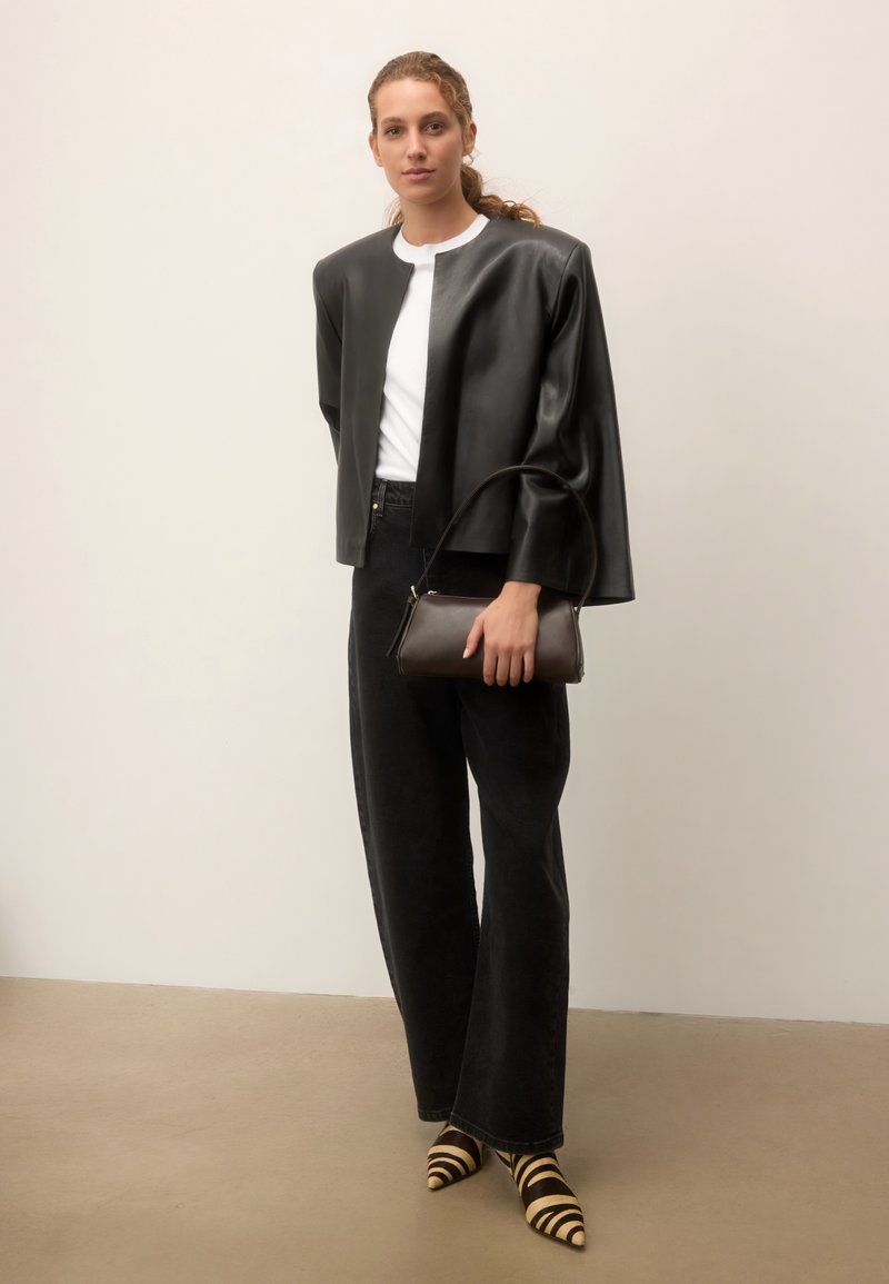 Femme aux cheveux attachés portant une veste en cuir noire, une chemise blanche, un pantalon noir, des chaussures rayées, tenant un petit sac à main sombre, debout à l'intérieur.