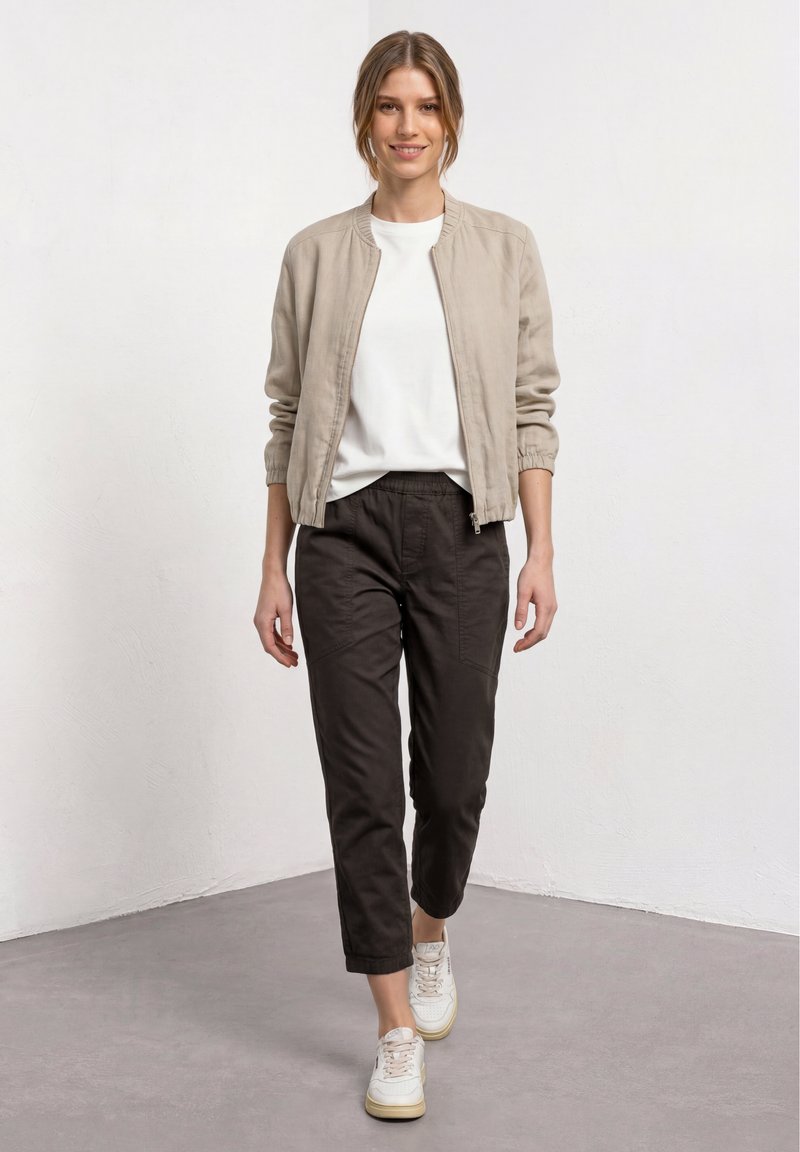 Jeune femme souriante marchant en avant portant une veste beige, une chemise blanche, un pantalon noir et des baskets blanches dans un environnement intérieur simple.