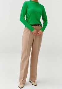 Personne portant un haut vert vif à manches longues, un pantalon beige taille haute avec une ceinture, et des chaussures crème pointues, debout les mains jointes.