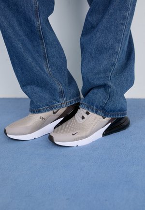 Persona que lleva zapatillas Nike beige y negras con suelas blancas y vaqueros azules, de pie sobre una alfombra azul claro.