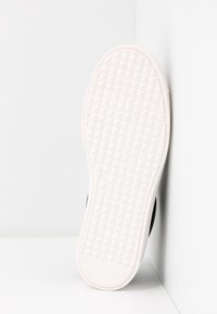 Semelle de chaussure en caoutchouc blanc avec motif de crampons en forme de grille reposant contre un mur d'angle blanc, taille de chaussure 37 visible sur la semelle.