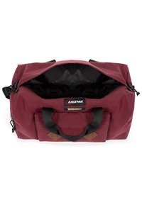 Borsa a duffel color bordeaux con manici neri, chiusura con zip e dettagli in pelle beige. Ampio interno nero con etichetta del marchio visibile all'interno.