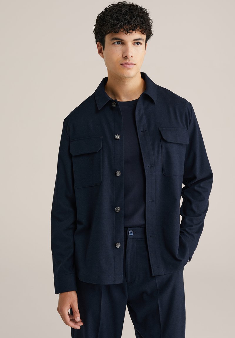 Marineblå skjorta med knappar och två bröstfickor, tillverkad av ett strukturerat tyg. Kombinerad med en matchande rundhalsad tröja och byxor.