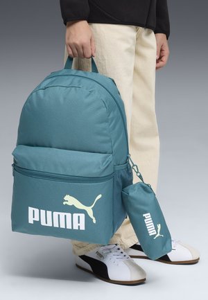 Persona sosteniendo una mochila Puma color azul verdoso con una bolsa a juego, vistiendo pantalones beige y zapatillas Puma blancas con detalles negros.