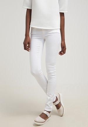 Person wearing slim white pants, white top, and off-white platform Mary Jane shoes with ankle straps, standing against a plain background.