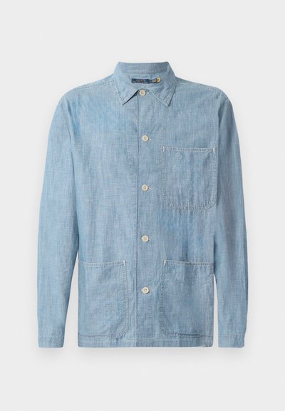 Svetlo modra srajca z gumbi iz denim tkanine, z ostrimi ovratniki, dvema sprednjima žepoma in bež gumbi. Teksturirana površina.