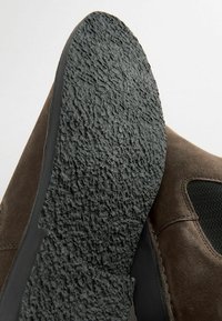 Chaussure en daim marron avec une semelle en caoutchouc noir texturé. Présente un dessus lisse et un panneau latéral à motifs. Accent sur l'adhérence et le contraste des matériaux.