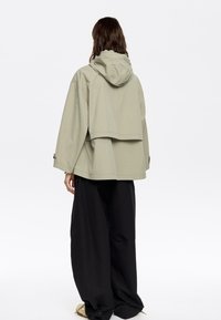 Giacca oversize verde chiaro con cappuccio, caratterizzata da un retro accorciato e maniche con bottoni, abbinata a pantaloni larghi neri.