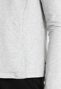 Světle šedá mikina s dlouhými rukávy, s jemnou strukturou a boční kapsou. Spodní lem je rovný s mírně elastickým zakončením.