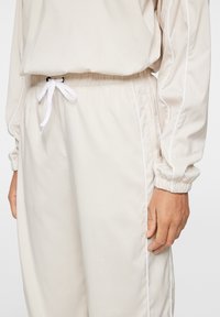 Beige träningsdräkt med elastisk midja och dragsko. Har vit kantning längs sidorna och elastiska ärmslut för en tapered passform.