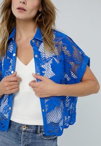 Blue lace top with floral pattern, short sleeves, and a shiny blue collar. Features a v-neck white undergarment and denim jeans.