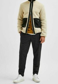 Fleecová bunda krémové barvy s černými doplňky a kapsami, v kombinaci s tmavě šedými kalhotami a bílými teniskami. Hladká textura, ležérní střih.