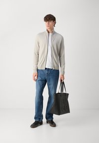 Lys grå zip-up cardigan over en hvit t-skjorte, kombinert med blå jeans. Holder en svart lær tote bag med preget "HUGO"-logo.