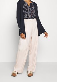 Granatowy kardigan z guzikami, bluzka w geometryczny wzór oraz jasnoróżowe spodnie o szerokich nogawkach. Całość dopełniają casualowe buty.