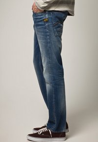 Siniset farkut suoran lahkeen istuvuudella, joissa on haalistunut pinta, kontrastiväriset ompeleet ja takataskun logo-merkki. Yhdistetty tummiin kenkiin.