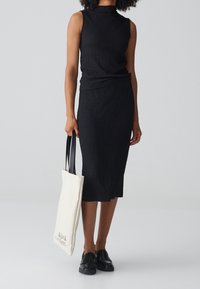 Haut sans manches noir et jupe longueur genoux en tissu texturé, accompagnés d'un sac fourre-tout crème avec des poignées noires et du texte imprimé.