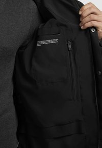 Sort vest med lynlåslommer, der har en glat, mat tekstur. Inkluderer et mærke med trykt hvid tekst. Tæt på visning af stoffet og designet.