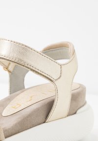 Béžové kožené sandály se zlatými akcenty, silnou bílou podrážkou a měkkou semišovou stélkou. Mají nastavitelné řemínky a polstrovaný design.